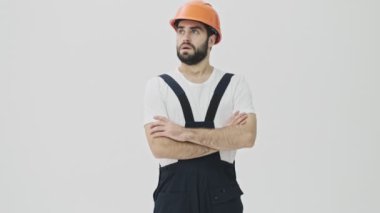 Kızgın, sakallı, genç bir inşaatçı miğferiyle beyaz duvar arkasında izole edilmiş.