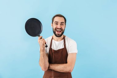 Heyecanlı genç sakallı aşçı önlük takıyor mavi arka planda tek başına duruyor, kızartma tavasını gösteriyor.