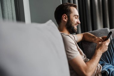 Evdeki kanepede cep telefonu kullanan yakışıklı, neşeli, mutlu ve pozitif bir adamın görüntüsü..