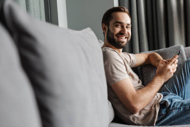 Evde, kanepede cep telefonuyla sohbet eden yakışıklı, pozitif bir genç resmi..