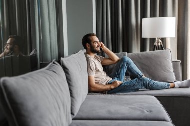 Evdeki kanepede akıllı telefondan konuşan yakışıklı, iyimser bir gencin görüntüsü..