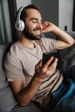 Evde, koltukta oturmuş kulaklıkla müzik dinleyen mutlu genç bir adamın görüntüsü..