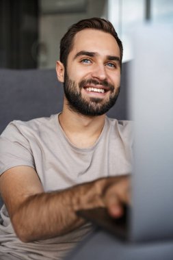 Evdeki kanepede dizüstü bilgisayar kullanan gülümseyen pozitif bir genç resmi..