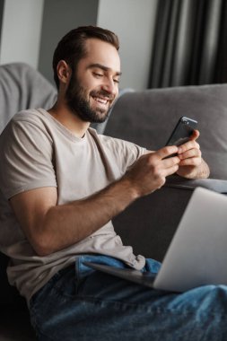 Evdeki kanepede bilgisayarla cep telefonuyla sohbet eden neşeli genç bir adamın görüntüsü..
