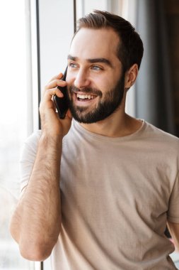 Evde cep telefonuyla konuşan gülümseyen pozitif bir genç resmi.