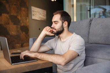 Çekici, ciddi, zeki bir genç adam oturma odasında yerde oturuyor ve dizüstü bilgisayarla çalışıyor.