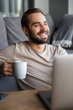 Çekici, mutlu, zeki bir genç adam oturma odasında yerde oturuyor, çalışıyor, dizüstü bilgisayar, kahve içiyor.