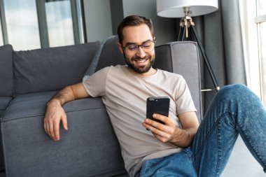 Çekici zeki genç adam oturma odasında yerde oturuyor, cep telefonu kullanıyor.