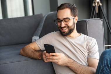 Çekici zeki genç adam oturma odasında yerde oturuyor, cep telefonu kullanıyor.