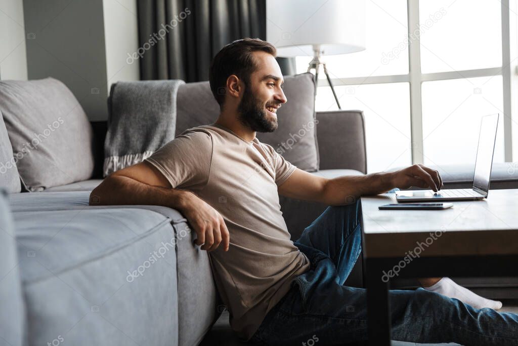 Image of a happy optimistic pleased young man indoors at home using laptop computer.