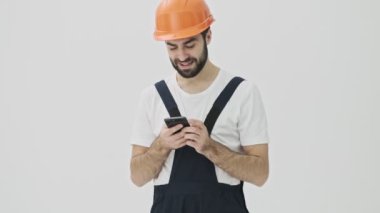 Neşeli iyimser genç sakallı adam miğferinde cep telefonu kullanarak beyaz duvar arkasında izole edilmiş.