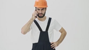 Stresli genç sakallı adam miğferiyle beyaz duvar arkasına izole edilmiş cep telefonuyla konuşuyor.