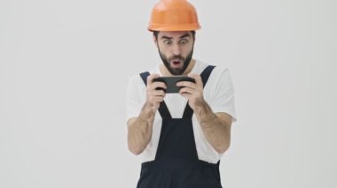 Heyecanlı, sakallı genç adam miğfer oyunlarında cep telefonuyla beyaz duvar arkasında izole edilmiş.