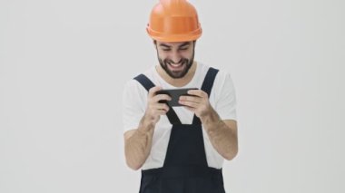 Yakışıklı, heyecanlı genç sakallı adam miğfer oyunlarında cep telefonuyla beyaz duvar arkasında izole edilmiş.