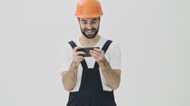 Heyecanlı, sakallı genç adam miğfer oyunlarında cep telefonuyla beyaz duvar arkasında izole edilmiş.