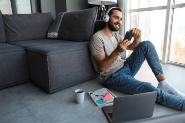 Evde oturup kulaklıkla müzik dinleyen mutlu ve iyimser bir gencin görüntüsü yerde oturup cep telefonuyla sohbet ediyor..
