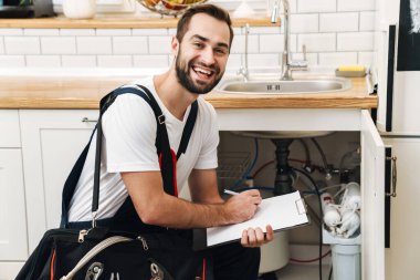 Apartman dairesinde çalışırken elinde ekipman ve dosya olan üniformalı beyaz bir tesisatçı resmi.