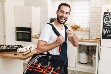 Apartman dairesinde çalışırken, elinde ekipman ve işaret parmağıyla çanta taşıyan, beyaz üniformalı genç bir tesisatçı resmi.