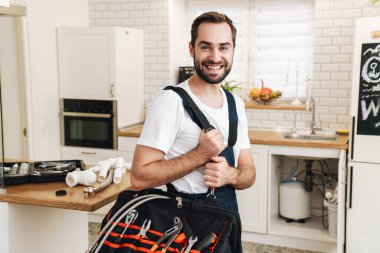 Kafkasyalı üniformalı genç bir tesisatçı resmi. Dairede çalışırken gülümseyen ve ekipmanlarla dolu bir çanta taşıyor.