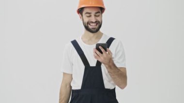 Mutlu, genç, sakallı bir adam miğferinde cep telefonu ve baş parmak hareketlerini gösteren bir miğferle beyaz duvar arkasında izole edilmiş.