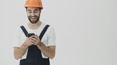 Pozitif, genç, sakallı adam miğferiyle beyaz duvar arkasına izole edilmiş cep telefonu kullanıyor..