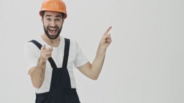 İyimser, mutlu, sakallı, genç bir inşaatçı beyaz duvar arkasına izole edilmiş miğferini gösteriyor..