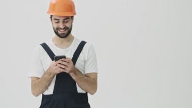 Pozitif genç sakallı adam miğferiyle beyaz duvar arkasına izole edilmiş cep telefonu kullanıyor.