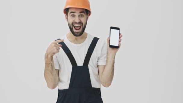 带着积极乐观的年轻胡子的建筑工人被隔离在白墙的背景下 头盔显示了手机的展示效果 — 图库视频影像
