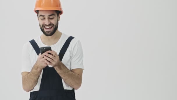 Pozitivní šťastný emocionální mladý vousatý muž stavitel izolované přes bílou zeď pozadí v přilbě zobrazující displej mobilního telefonu.