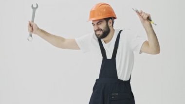İyimser genç sakallı inşaatçı beyaz duvar arkasında ingiliz anahtarıyla dans ediyor.