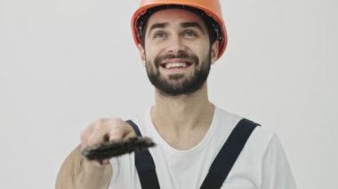 Mutlu, sakallı, genç bir inşaatçı beyaz duvar arkasında, fırçalı bir kaskla izole edilmiş.