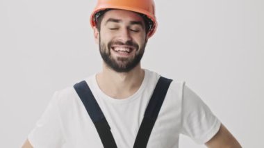 Mutlu, genç, sakallı adam miğferiyle beyaz duvar arkasına izole edilmiş, başparmak işareti yapıyor.