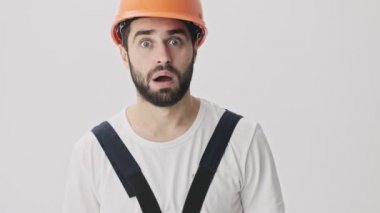 Şok olmuş genç sakallı inşaatçı kaskla beyaz duvar arkasında izole edilmiş.