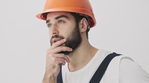 认为年轻的留着胡子的建筑工人戴着头盔 被隔离在白墙的背景之外 — 图库视频影像
