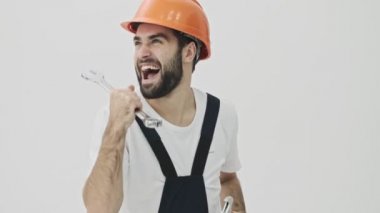 İyimser genç sakallı inşaatçı beyaz duvar arkasında ingiliz anahtarıyla dans ediyor.
