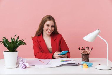 Neşeli genç bir kadının görüntüsü kapalı bir masada pembe duvar arkasında izole bir şekilde cep telefonuyla sohbet ediyor..