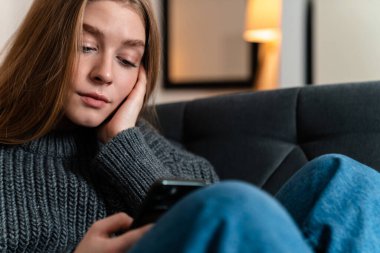 Evde cep telefonu kullanan konsantre olmuş genç bir kadının görüntüsü.