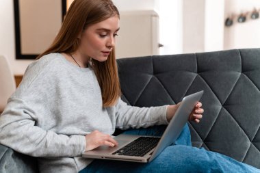 Evdeki kanepede dizüstü bilgisayar kullanan konsantre olmuş genç bir kadının fotoğrafı..