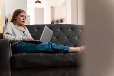 Evdeki kanepede dizüstü bilgisayar kullanan üzgün ve korkmuş genç bir kadının fotoğrafı..