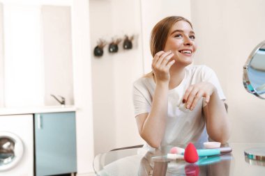 Evde, yatak odasında, aynayla makyaj yapan mutlu ve konsantre genç bir kız resmi.