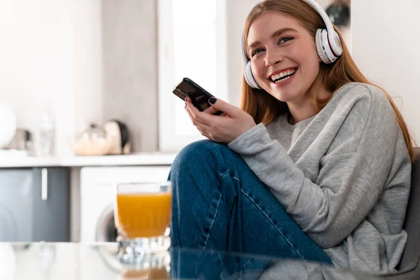 照片中 乐观的年轻女子在家中用手机听音乐 带耳机 — 图库照片
