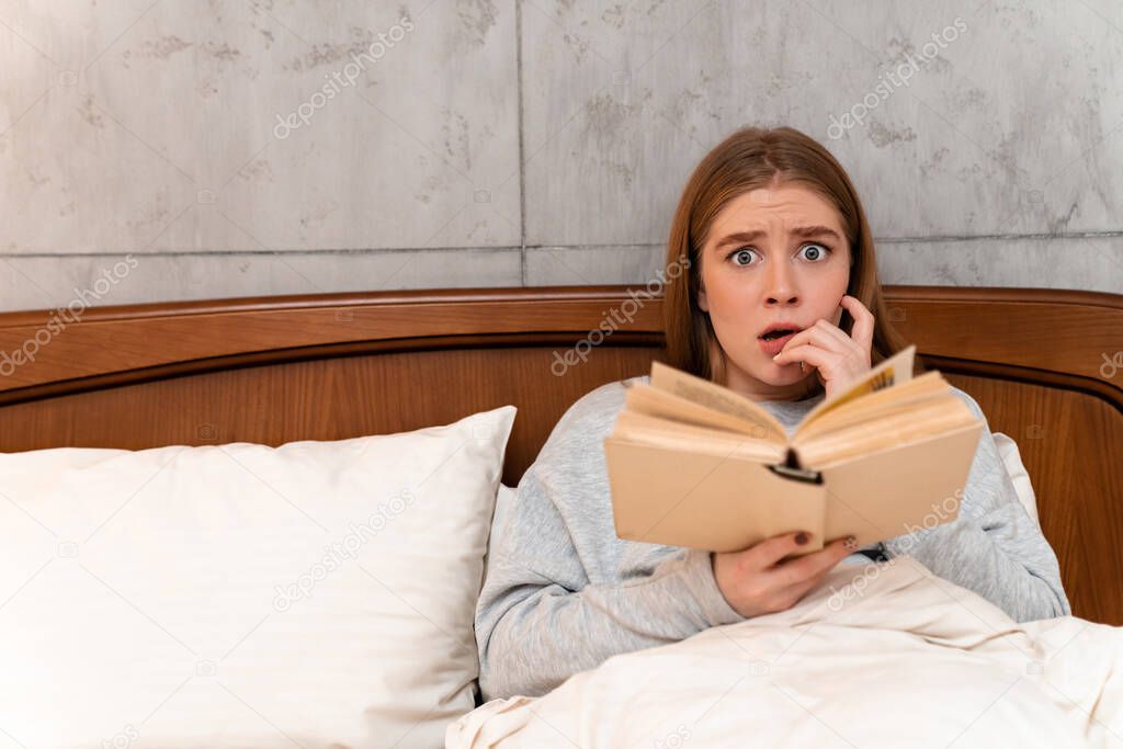 Photo of a shocked scared pretty young woman indoors at home in bed reading book.