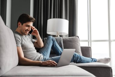 Oturma odasındaki kanepede oturmuş dizüstü bilgisayar kullanırken cep telefonuyla konuşan ve sıradan kıyafetler giyen çekici, gülümseyen genç bir adamın portresi.