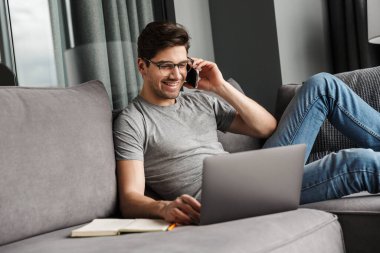 Oturma odasındaki kanepede oturmuş dizüstü bilgisayar kullanırken cep telefonuyla konuşan ve sıradan kıyafetler giyen çekici, gülümseyen genç bir adamın portresi.