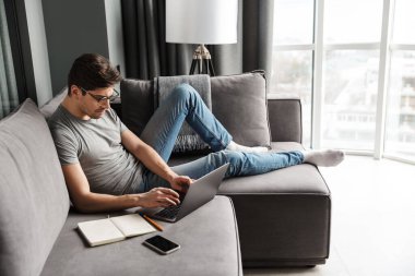 Oturma odasındaki kanepede oturup dizüstü bilgisayar kullanan, gündelik giysiler giyen çekici, ciddi ve sakallı bir adamın portresi.