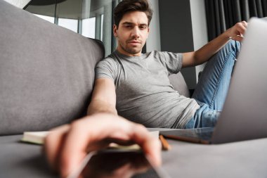 Oturma odasındaki bir kanepede oturan, dizüstü bilgisayarla uğraşırken elinde boş cep telefonu olan, ciddi ve ciddi bir sakallı genç adam.