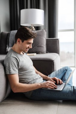 Çekici gülümseyen sakallı genç bir adamın portresi gündelik giysiler giymiş oturma odasındaki koltukta oturmuş dizüstü bilgisayar kullanıyor.