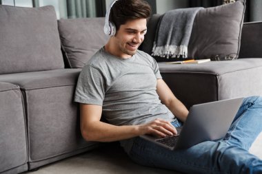 Oturma odasındaki kanepede oturmuş, kablosuz kulaklık takmış, dizüstü bilgisayar kullanan, gündelik giysiler giyen çekici, genç sakallı bir adamın portresi.