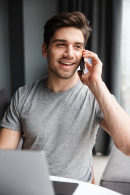 Yakışıklı, sakallı, sıradan kıyafetler giyen, evde otururken dizüstü bilgisayar kullanan bir adam.