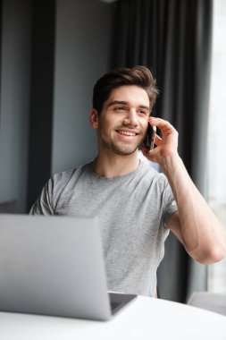 Yakışıklı, sakallı, sıradan kıyafetler giyen, evde oturmuş cep telefonuyla konuşurken dizüstü bilgisayar kullanan bir adam.